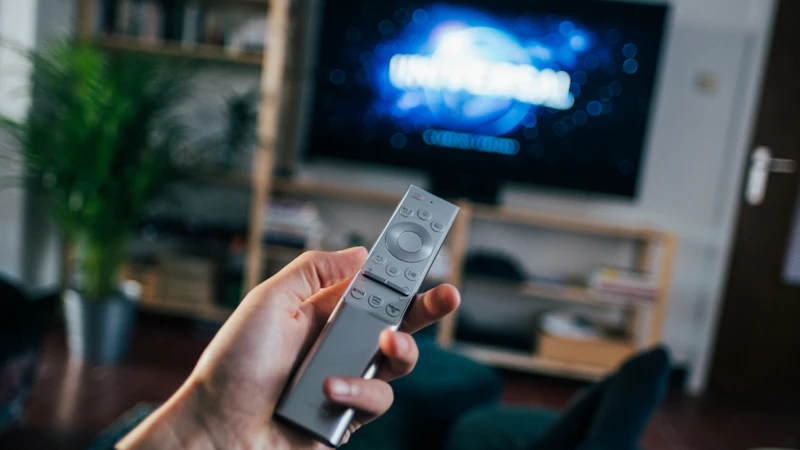 Hand hält eine Fernbedienung vor einem eingeschalteten Fernseher im Wohnzimmer
